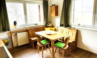 A bright kitchen with a wooden dining area and green chairs. Large windows provide plenty of daylight.