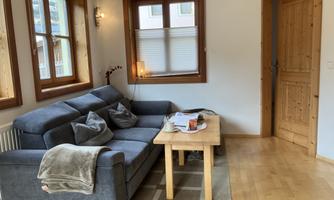 A cozy living room with a blue sofa and a wooden table. Large windows let in plenty of light and the walls are painted in warm colors.