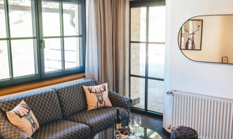 A cozy living room with a sofa, a glass table, and a small stool. Large windows provide plenty of natural light.