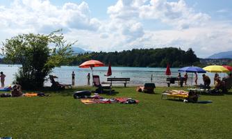 A beautiful lake with grassy shores, sun loungers, and colorful beach umbrellas. People are enjoying the water and the sunny atmosphere.