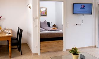 A modern, bright room with a dining table and chairs. In the background, there is a bedroom with a bed and a television.