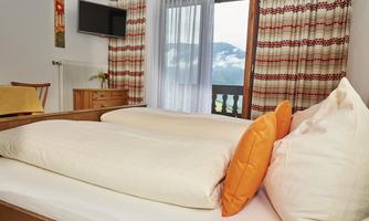 A cozy bedroom with two beds and orange pillows. Large windows offer a view of the mountains, and light streams into the room through the window.