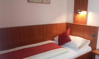 A simple single room with a wooden frame, a comfortable bed, and a red pillow. The walls are light, and there is a wall lamp for extra light.