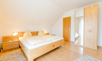 A bright bedroom with a double bed and two nightstands. The room features a large mirror and a warm wooden floor.