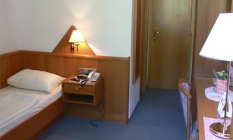 A simple hotel room with a single bed, a nightstand, and a lamp. Next to the bed is a table with a chair and a small decoration.
