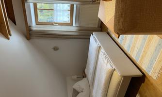 A cozy bedroom with a double bed and a window that lets in daylight. The floor is covered with a light carpet, and there is a small decorative corner.