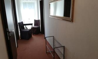 A bright hallway with carpet and a window that lets in daylight. On the left side, there are two chairs and a table, and a mirror is hanging on the wall.