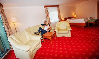 A cozy hotel room with a red carpet, beige furniture, and large windows. A person is sitting comfortably in an armchair.