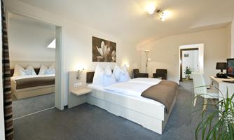 A modern bedroom with a double bed and stylish pillows. In the background, there is a comfortable seating area and an additional bedroom.