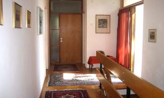 A bright hallway with wooden details and colorful carpets. Pictures hang on the walls and a large window lets in natural light.