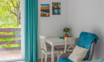 A cozy room with a bed, an armchair, and a table. The walls are white, and there are light curtains as well as some pictures on the wall.