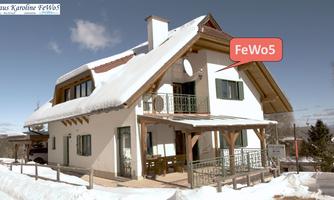 A cozy holiday home with wooden elements and a balcony. The surroundings are covered in snow and the sky is clear.