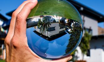 A hand holds a crystal ball that reflects a green meadow and a building. The sky is clear and blue.