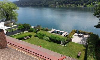 A peaceful landscape with a clear lake, surrounded by green mountains. In the foreground, there is a well-kept garden with a pool and seating areas.