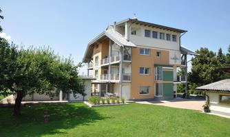 A modern house with multiple floors and large windows stands in a well-kept garden. In the background, some trees and a carport can be seen.