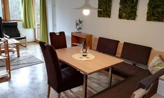 A cozy dining area with a wooden tray and comfortable chairs. In the background, there are large windows and decorative plant wall art.