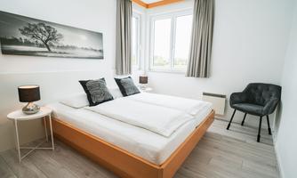 A modern bedroom with a large double bed and stylish pillows. The room has large windows and elegant furniture.