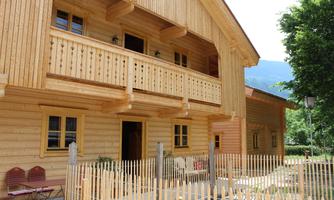 A traditional wooden house with a balcony and large windows. The surrounding landscape is green and inviting.