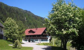 A picturesque house with a red roof in a green landscape. Surrounded by trees and mountains under a clear blue sky.