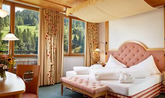 A cozy hotel room with a large bed and blooming curtains. Large windows offer a view of the green landscape.