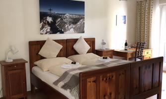 A cozy bedroom with a wooden bed and two pillows. A picture of a mountain landscape hangs on the wall, and there is a small workspace with a desk.