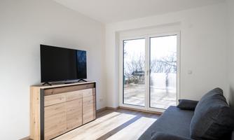 A modern living room with a large window and a beautiful view. A television stands on a wooden cabinet next to a gray sofa.
