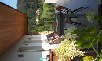 A sunny terrace with a table and chairs, surrounded by green plants. In the background, hills and an idyllic village can be seen.