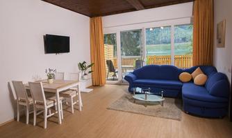 A bright living room with a blue sofa and a dining table. Large windows offer a view of the outdoor landscape.