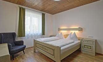 A cozy bedroom with a double bed and two pillows. There is an armchair and windows with green curtains that provide daylight.