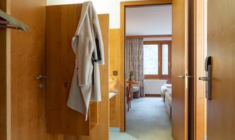 A bright hallway with wooden details and two open doors. A cozy room with windows is visible in the background.