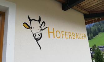 A stylized cow motif on a wall with the inscription "Hoferbauer". The surroundings show a green slope in the background.