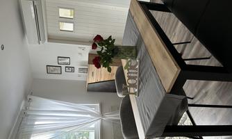 A cozy dining area with a wooden table, a vase with red roses, and a sign saying "Home". In the background, there are windows and a modern room visible.