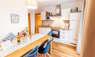 A modern kitchen with a wooden dining table and blue chairs. The room is warmly lit and well-furnished.