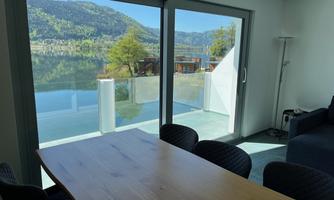 A modern room with a large table and chairs that offers a view of a tranquil lake and mountains. Large windows allow plenty of light in.