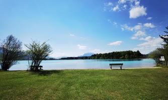 A quiet lake with clear blue water and green meadows. In the foreground, two benches and some trees are visible.