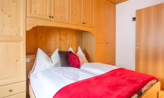 A cozy bedroom with a wooden bed and red pillows. The walls are made of light wood, and there is a door in the background.