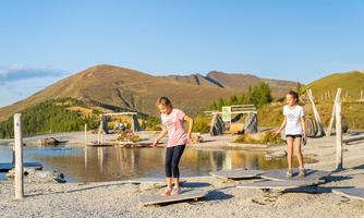 Two girls are standing on wooden platforms at the shore of a clear lake. In the background, gentle hills and trees can be seen.