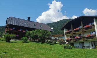 A picturesque holiday home with a large garden and colorful flowers on the balconies. In the background, green hills and a blue sky with clouds can be seen.