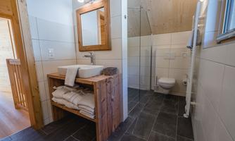 A modern bathroom with an open shower and a wooden sink. Towels are neatly stacked and a large mirror hangs on the wall.
