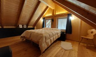A cozy attic room with wooden paneling and sloped walls. There is a bed, windows with curtains, and a light wooden chair.