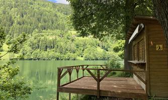 A cozy cabin by the lake, surrounded by lush greenery. In the background, gentle hills rise beneath a clear sky.