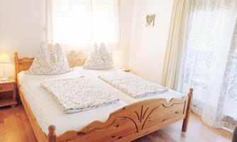 A cozy bedroom with a wooden double bed and large pillows. The windows are decorated with curtains, and the light shines in warmly.