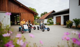 A family is riding three-wheeled vehicles on a farm. In the foreground, flowers can be seen, creating a friendly atmosphere.