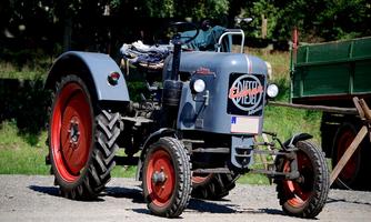 Ein klassischer Traktor mit grauer Karosserie und roten Rädern steht auf einem Schotterweg. Im Hintergrund ist ein grüner Anhänger zu sehen.