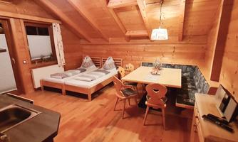 A cozy room with wooden walls and a large table. In the background, a bed and a kitchen area can be seen.