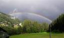 A rainbow stretches over a green landscape with hills and trees. The sky is cloudy, which gives the scene a dramatic atmosphere.