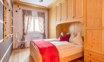 A cozy bedroom with a wooden bed and red accents. The walls are painted in neutral colors and the window lets in plenty of daylight.
