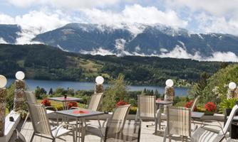 A picturesque landscape with mountains and a lake. On the terrace, there are tables and chairs, surrounded by flowers.