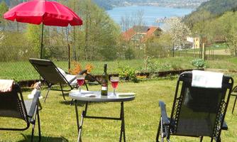 A beautiful garden with loungers and a red sun umbrella. On a table, there are two glasses of wine and a bottle, while there's a view of the lake.