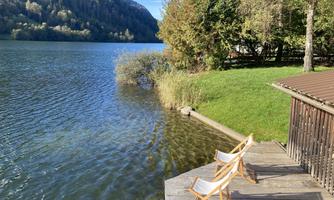 A tranquil lakeside landscape with two folding chairs on a wooden dock. The surroundings are green with trees and mountains in the background.